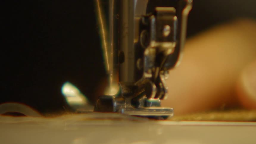 The metal needle of a sewing machine pumps rapidly in a rhythmic blur driving stitches into the fabric as the presser foot guides the material under warm golden light