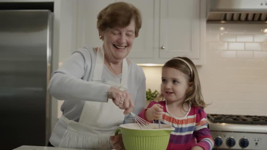 Older woman in a kitchen with a child and both of them are stirring something in the same bowl