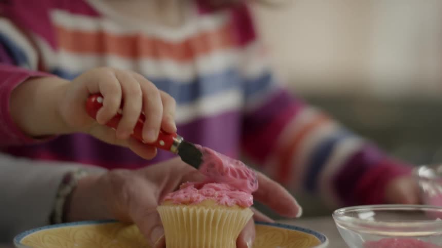 Child spreads pink frosting on a cupcake