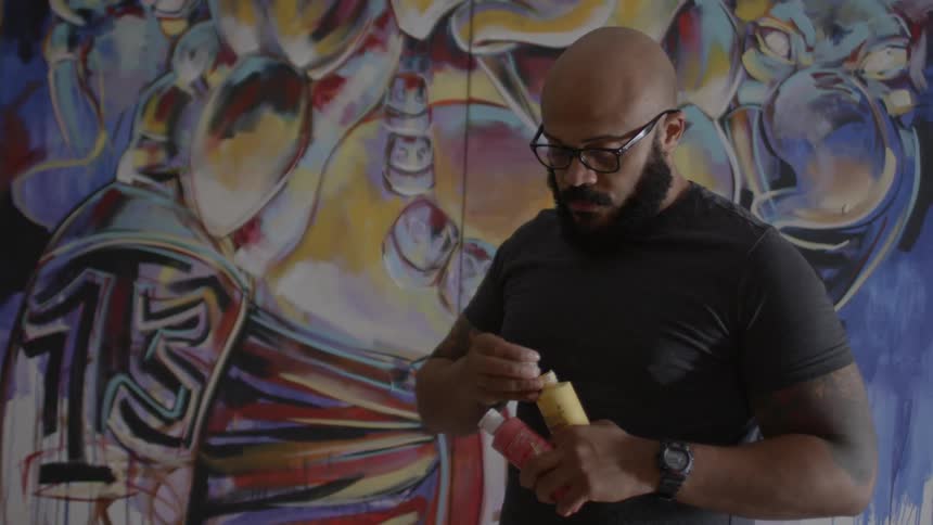 Bearded man removing the cap off a yellow bottle before a wall that has graffiti-like painting on it