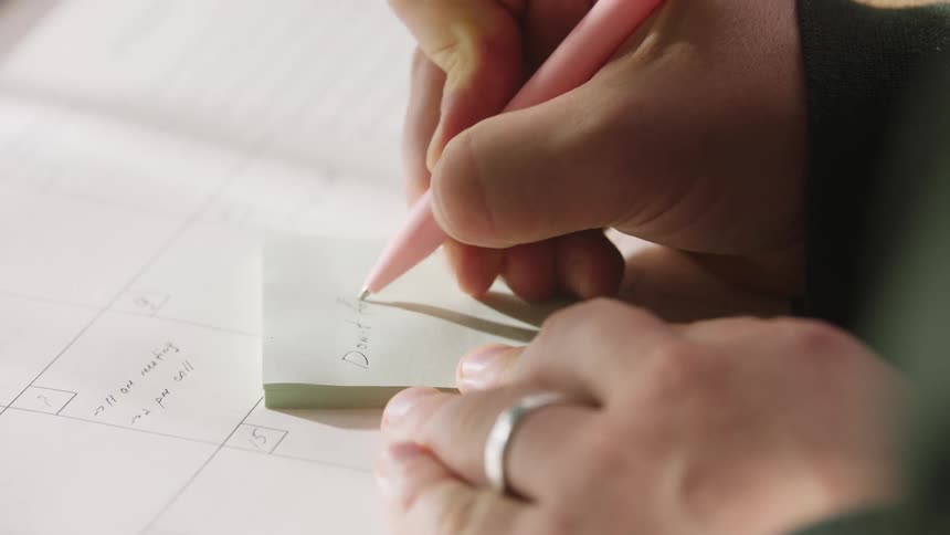 A person uses a pink pen to write dont forget on a sticky note placed on top of a calendar