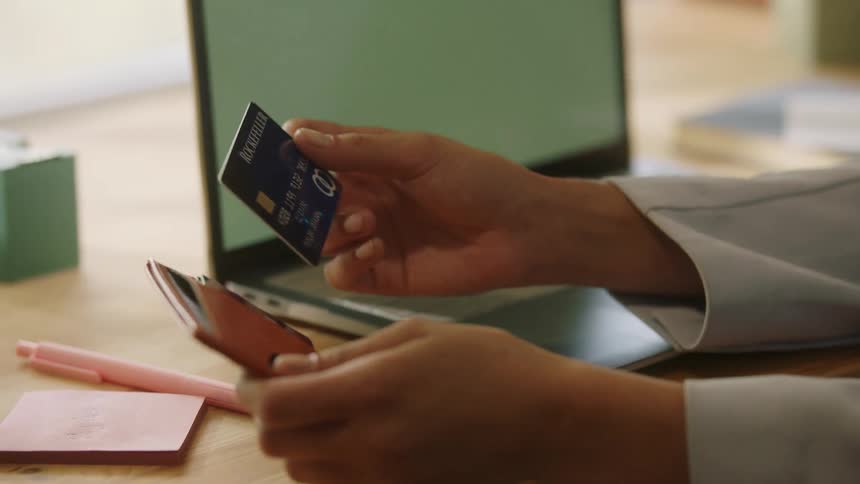 Person pulls dark card from brown wallet on desk with laptop and stationery items