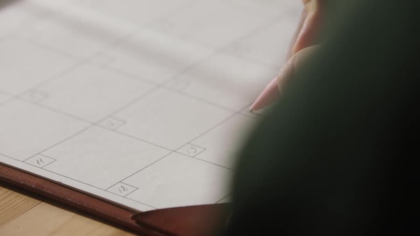 Hand writes with a pencil on a calendar page with organized dates