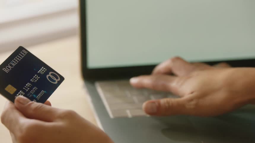 Hand holds blue card near laptop on wooden surface with wallet beside it
