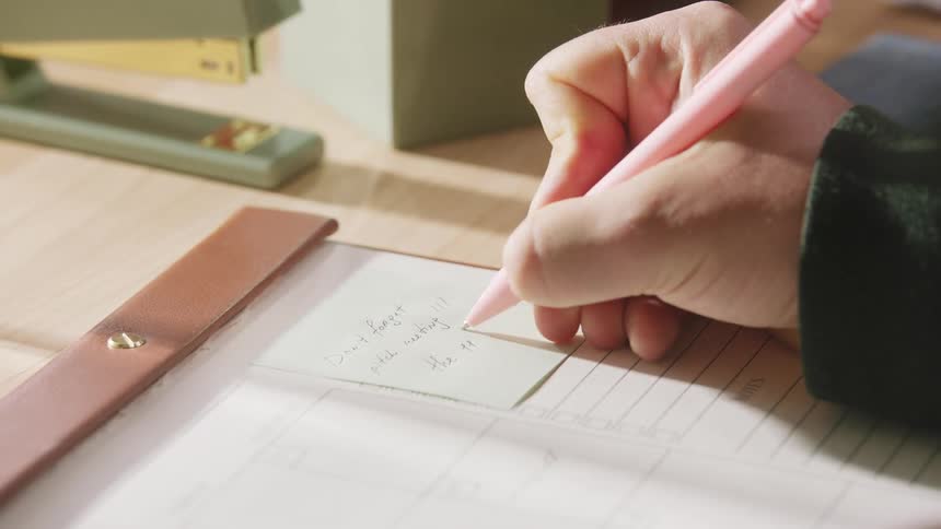 An adult hand writes on a sticky note with a pink ink pen