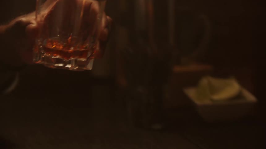 A closeup shot focuses on a faceted glass tumbler filled with a dark amber liquid on a bar counter which is then picked up by a hand and lifted toward a mans mouth in a dimly lit setting