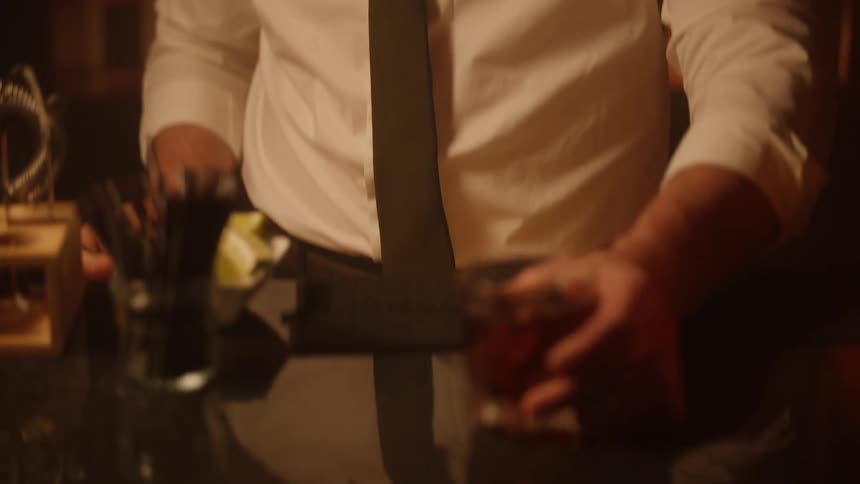 A bartender stirs a dark cocktail in a rocks glass with a long spoon then places the finished drink on the reflective bar top in a warmly lit room