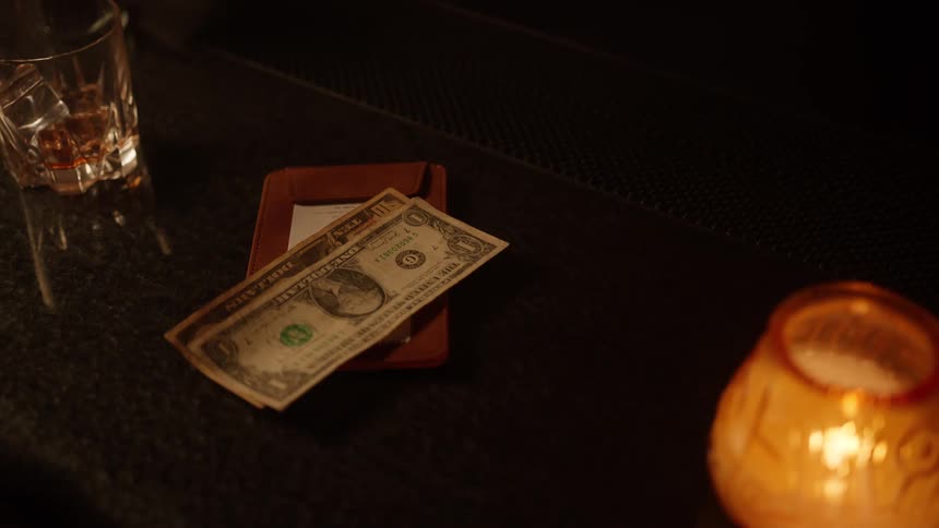 A closeup shot focuses on a brown leather check holder containing a receipt on a dark table where a us dollar bill is briefly placed on top before a persons hand picks up the cash and the check holder