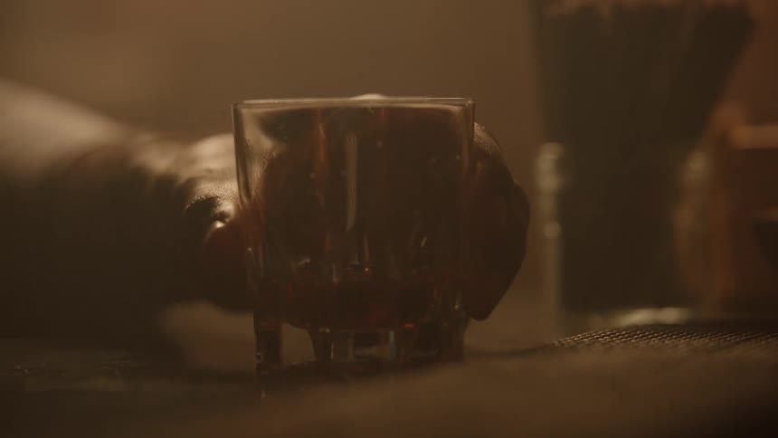 A hand setting a short faceted glass tumbler which holds some whiskey down onto a dark surface in a warmly lit hazy bar environment and a second hand takes the glass
