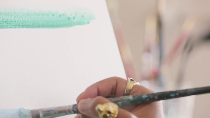 A hand adorned with gold rings paints a smooth light blue stroke across a white canvas