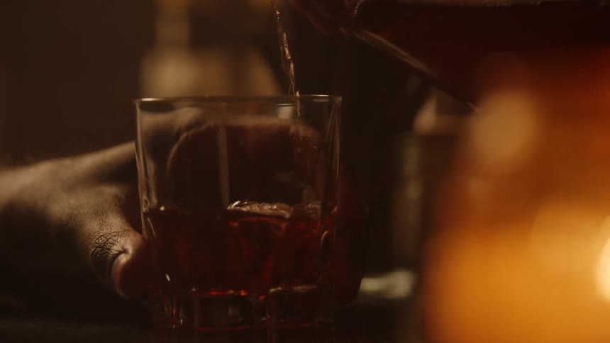 A closeup shot shows a hand gripping a faceted glass on a dark bar counter as a stream of dark amber liquid is steadily poured from above filling the glass in a warmly lit intimate bar setting