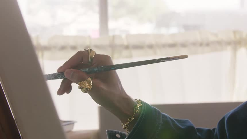 Hand with gold rings holding brush in front of canvas with blue paint traces on fingers