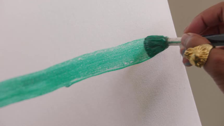 Brush applying green paint on white surface with visible gold rings on hand