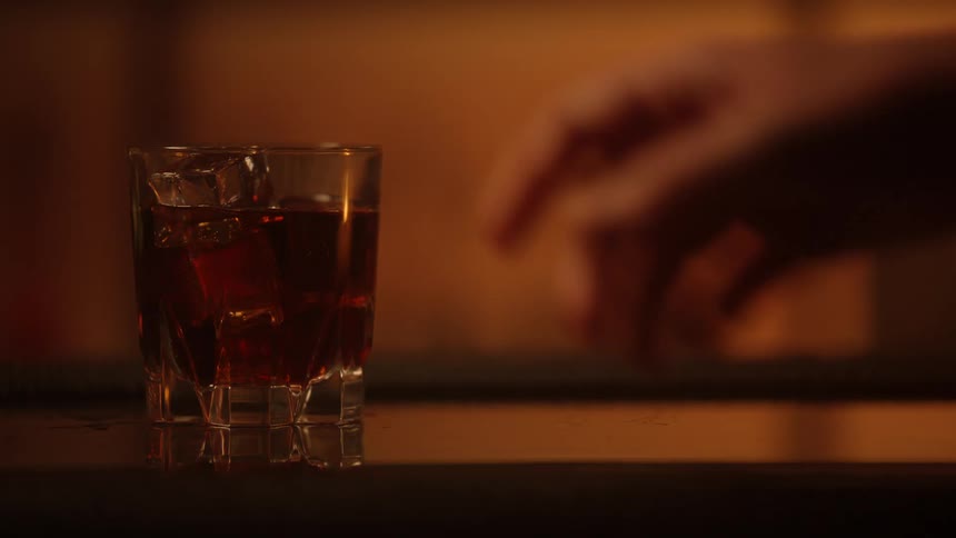 Hand holds glass with amber drink and ice in warmly lit setting