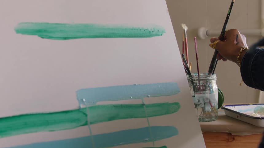 A hand cleans a paintbrush by dipping it into a mason jar of water and wiping the excess against the rim