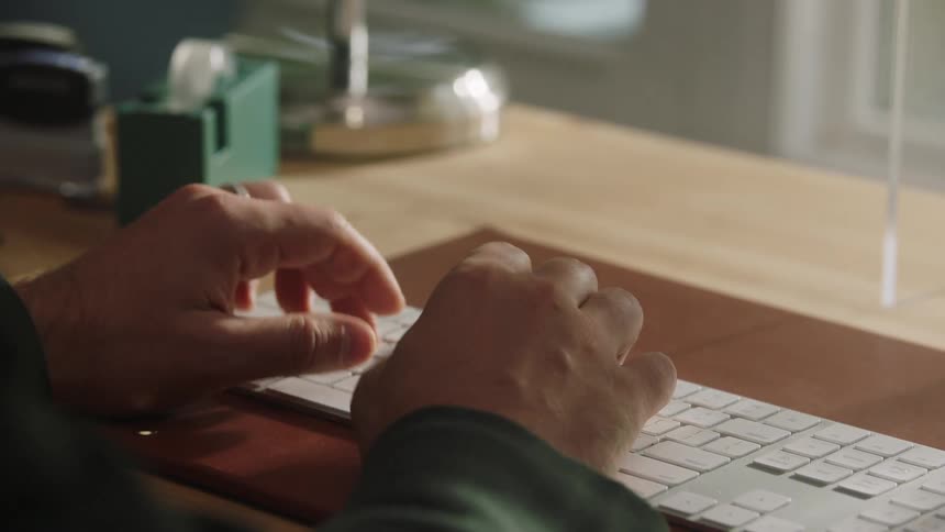 A closeup of hands typing on a white keyboard at a wooden desk indoors