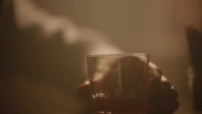 A persons hand reaches for and lifts a faceted glass tumbler containing an orangecolored beverage bringing the drink up to the mouth for a sip in a dimly lit atmospheric setting