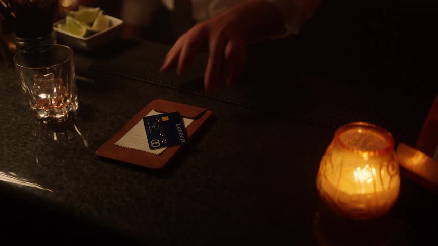 A customer places a credit card on a bill holder at a candlelit bar and the bartender collects it to process the transaction