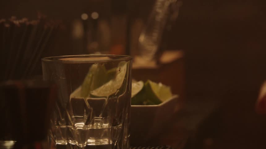 A bartender holds a silver cocktail shaker straining a clear liquid from the shaker into a heavy faceted glass tumbler which already contains a lime wedge on a dark bar counter