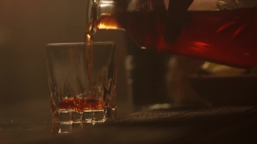 A hand places a cutglass tumbler onto a bar surface in dim lighting a reddishbrown liquid likely whiskey or a similar spirit is poured into the glass from a bottle