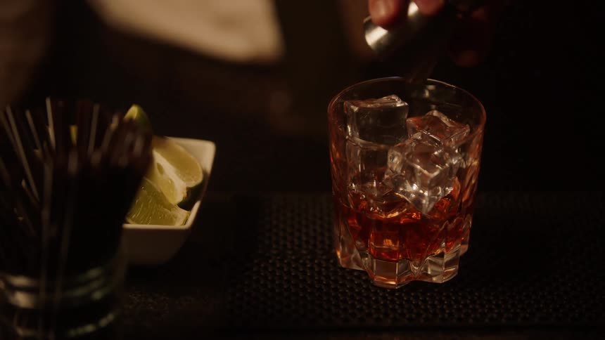 A bartender wearing a white shirt and dark tie uses a jigger to measure and pour a dark amber liquor into a faceted glass filled with ice then adds a splash of a secondary component to complete the cocktail on a dark bar counter