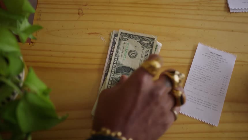 A black womans hand decorated with gold rings and bangles places cash on a desk for payment as the cashiers hand reaches forward to collect it with a receipt lying on the desk