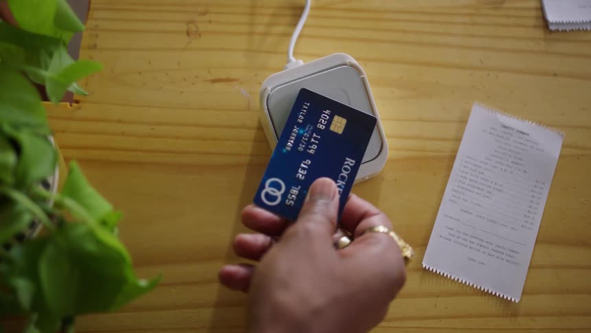 A human hand using a blue credit card to approach a payment device next to a receipt