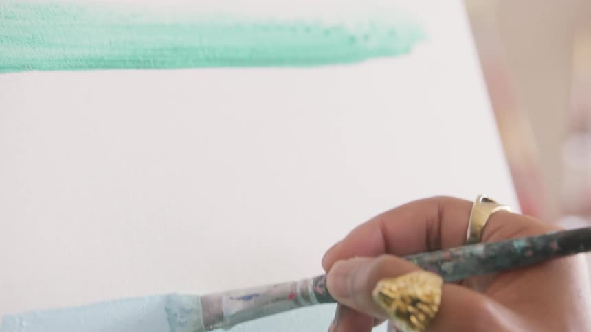 A hand wearing prominent gold rings holds a paintbrush and applies a broad stroke of light blue paint across a white surface this stroke is laid just above a previously painted mint green line