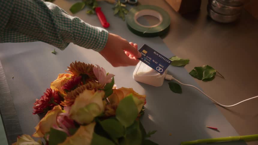 A persons hand using a blue credit card to pay at a contactless payment machine next to some flowers