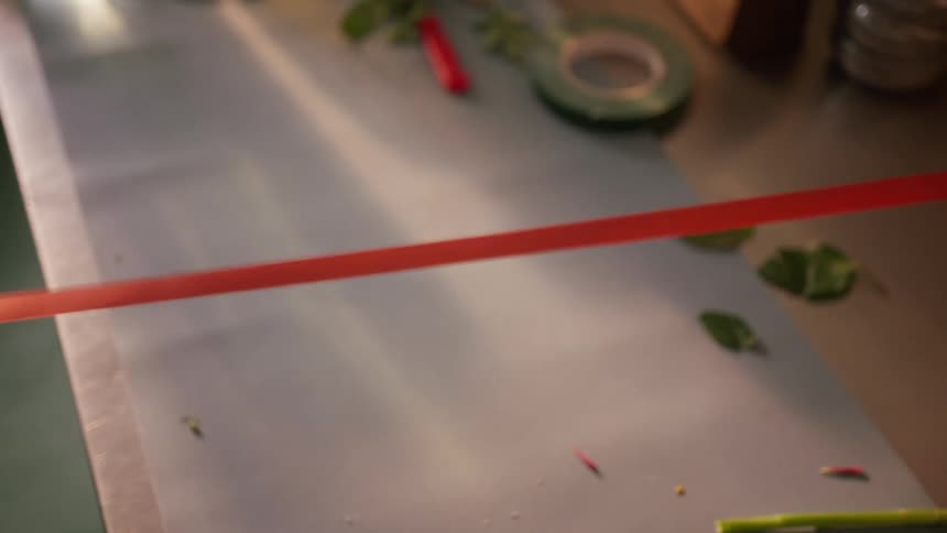 A persons hand holds a pair of small scissors to cut a long thin red ribbon that is stretched across a large lightcolored surface which also has some loose green leaves and a roll of green tape in the blurred background