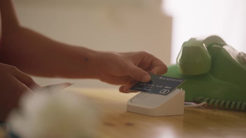 A person inserts a credit card into a small reader beside a green rotary phone on a wooden counter