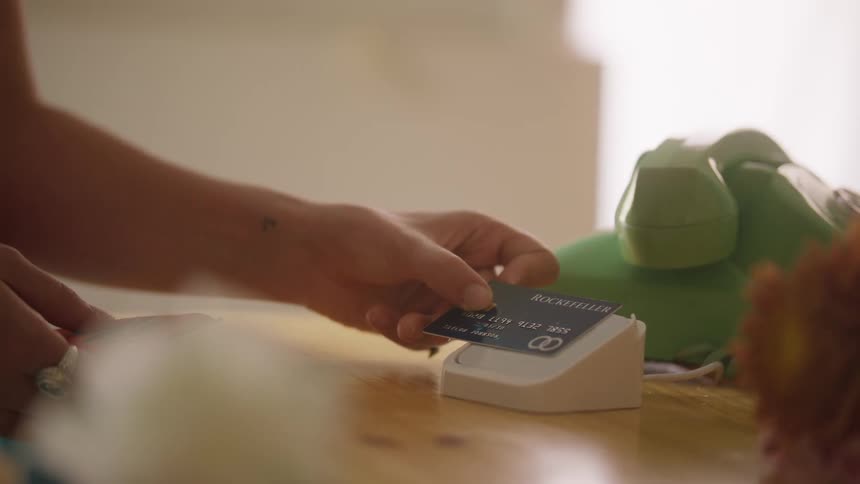 A customer places a single flower onto the counter while simultaneously tapping a black credit card against the white payment terminal near a vintage green rotary phone