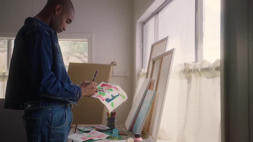 A young man with a shaved head wearing a denim jacket and jeans stands in a brightly lit room near a large window intently focusing as he paints on a small piece of paper covered with abstract colors