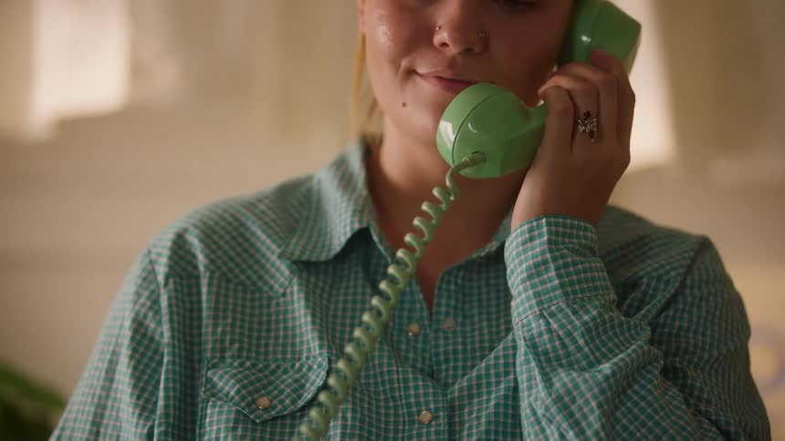 A woman picks up a vintage green telephone and smiles warmly as she brings the receiver to her ear to answer a call in a softly lit room