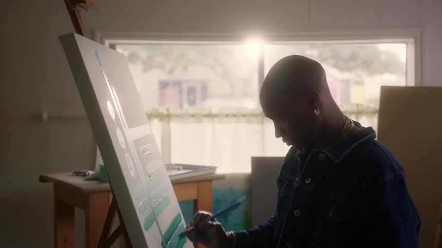 A painter leans over his canvas adding detailed green brushstrokes to his work as soft light streams in through a large studio window