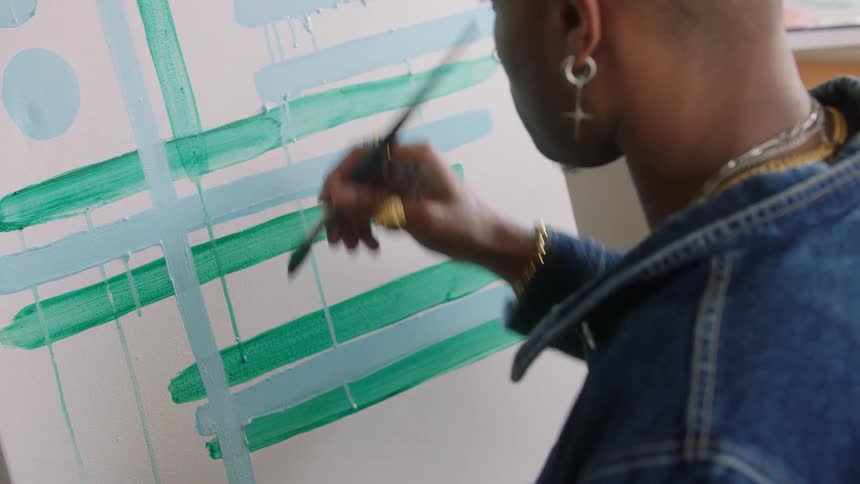 Seen from over the shoulder an artist methodically paints bold green and blue lines adding layers to an abstract piece on a large canvas