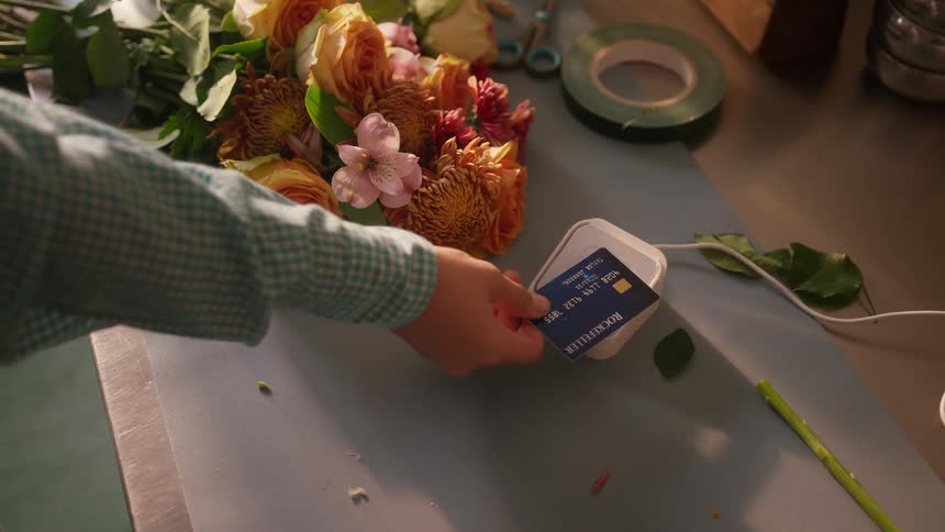 A person taps a credit card on a white reader placed on a table next to a bouquet of flowers