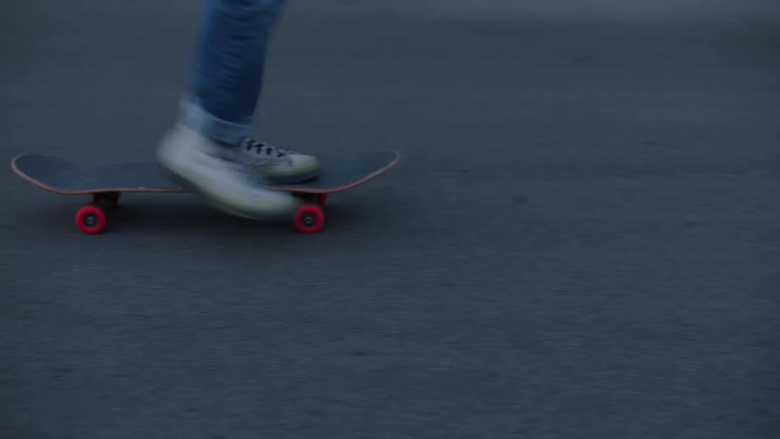 A person wearing jeans rides a skateboard across an asphalt surface at midday dressed in white sneakers