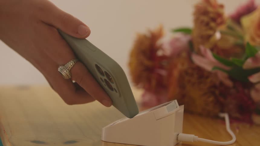 A woman makes a quick contactless payment by tapping her phone on a card reader with a vibrant bouquet of flowers blurred in the background