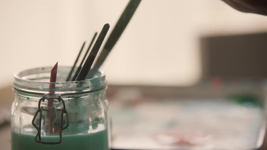 A closeup shot focuses on a glass jar containing murky bluegreen wash water and several paintbrushes as a hand wearing gold jewelry dips or removes a brush from the jar with a painters tray visible nearby