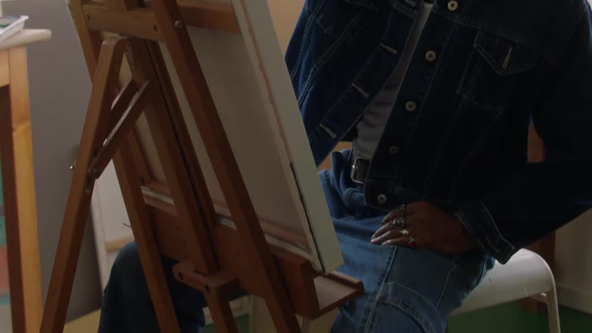 Painted papers lie scattered on the floor beneath a man in a denim jacket who sits painting at an easel