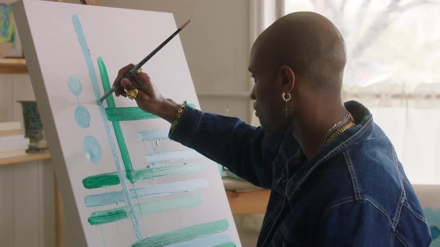 An artist paints abstract teal and green shapes on a white canvas indoors