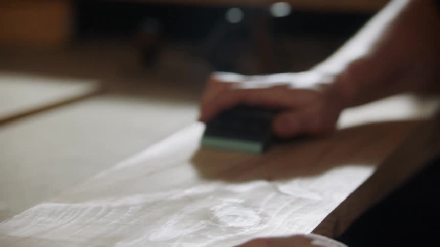 The video shows a closeup of a person sanding a piece of wood the motion is repetitive moving a sanding block back and forth along the grain of the wood