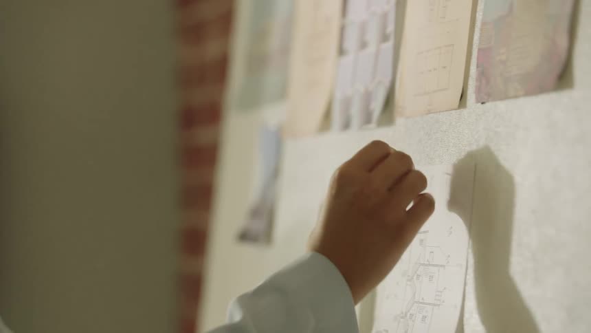 A man wearing a lightcolored jacket is seen picking up a small architectural drawing from a wooden shelf and then using his hands to pin the plan onto a wall already partially covered with other similar documents