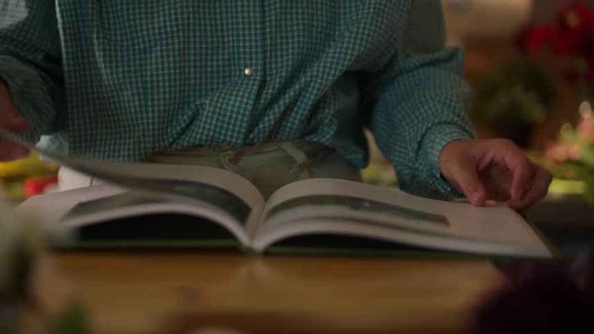 Person reading illustrated book on wooden table in plantfilled setting