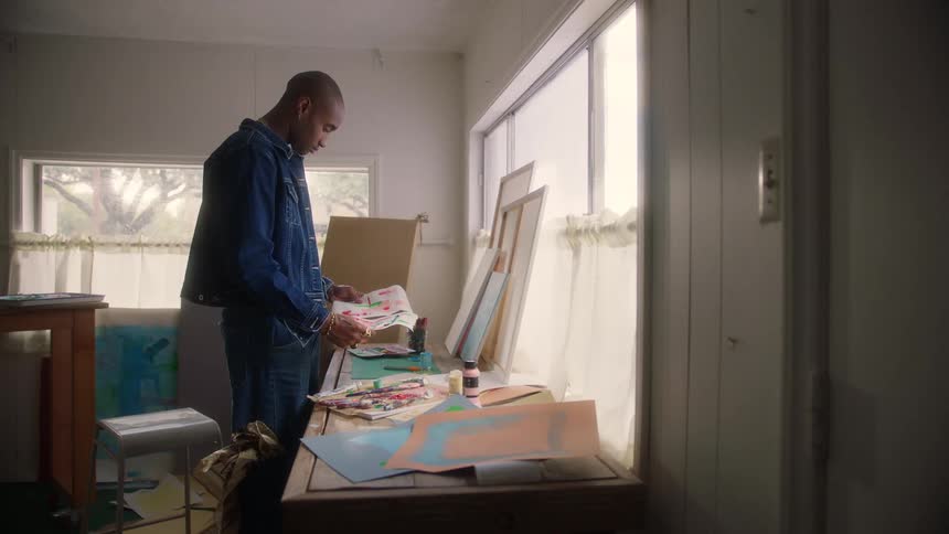 A man in a denim jacket stands at a sunlit table sorting through various paintings on paper