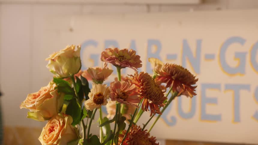 Person wearing a green plaid shirt arranges a small bouquet of orange and yellow flowers in front of a colorful sign