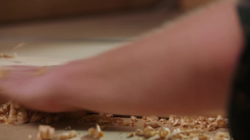 A closeup shot focuses on a piece of lightcolored wood covered in numerous thin curled wood shavings which are then quickly swept clear by a hand across the surface