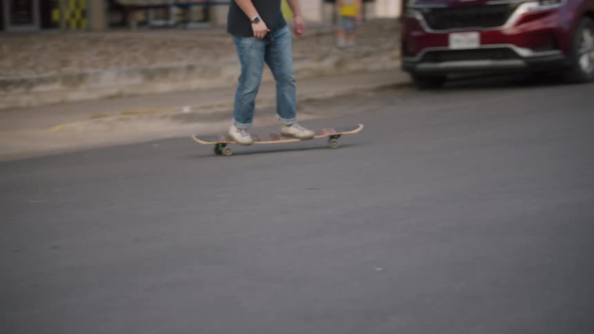 A person in jeans rides a longboard down a quiet street pushing off once to gain speed before cruising along the smooth pavement