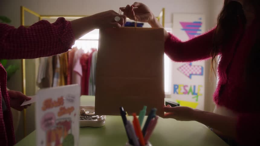 In a bright clothing or resale shop a woman wearing a bright pink fuzzy sweater hands a tall brown paper shopping bag across a counter to another person who is simultaneously exchanging a credit cardfor the transaction